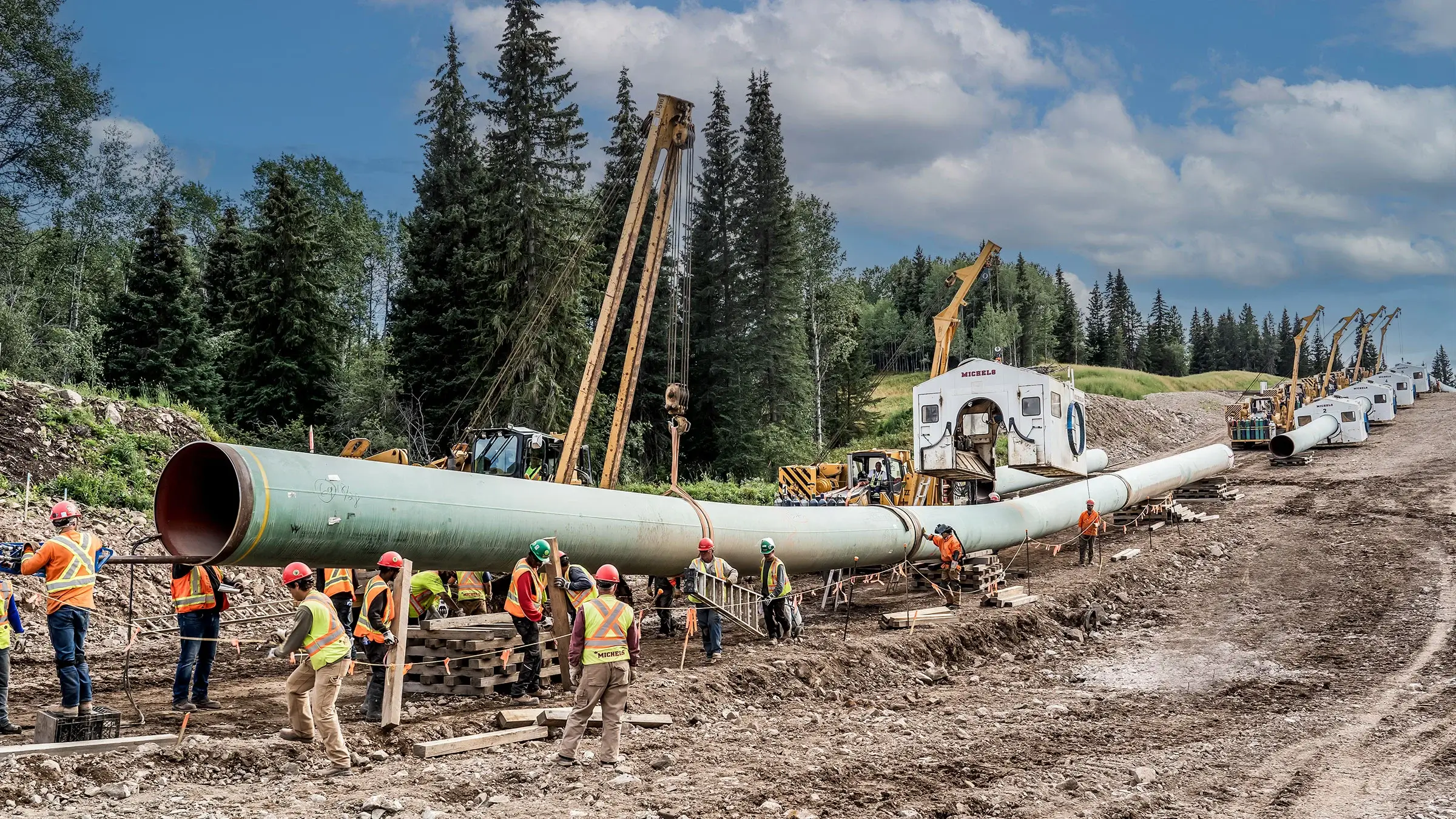 Welders work along a long suspended pipeline in the forest.